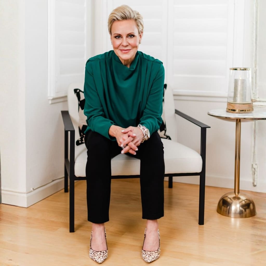 A woman with short blonde hair sits on a white chair, wearing a green blouse, black pants, and animal print shoes—hands clasped—in a bright Los Angeles room with white shutters, embodying the chic touch of AC.Styles' personal shopping service.